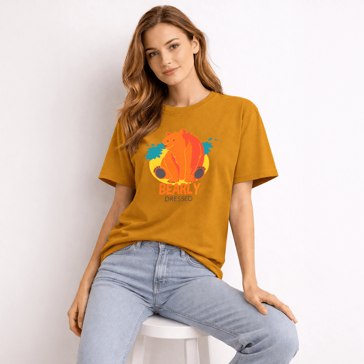Woman wearing a mustard yellow t-shirt with a colorful bear graphic and text, sitting on a white stool against a light background.